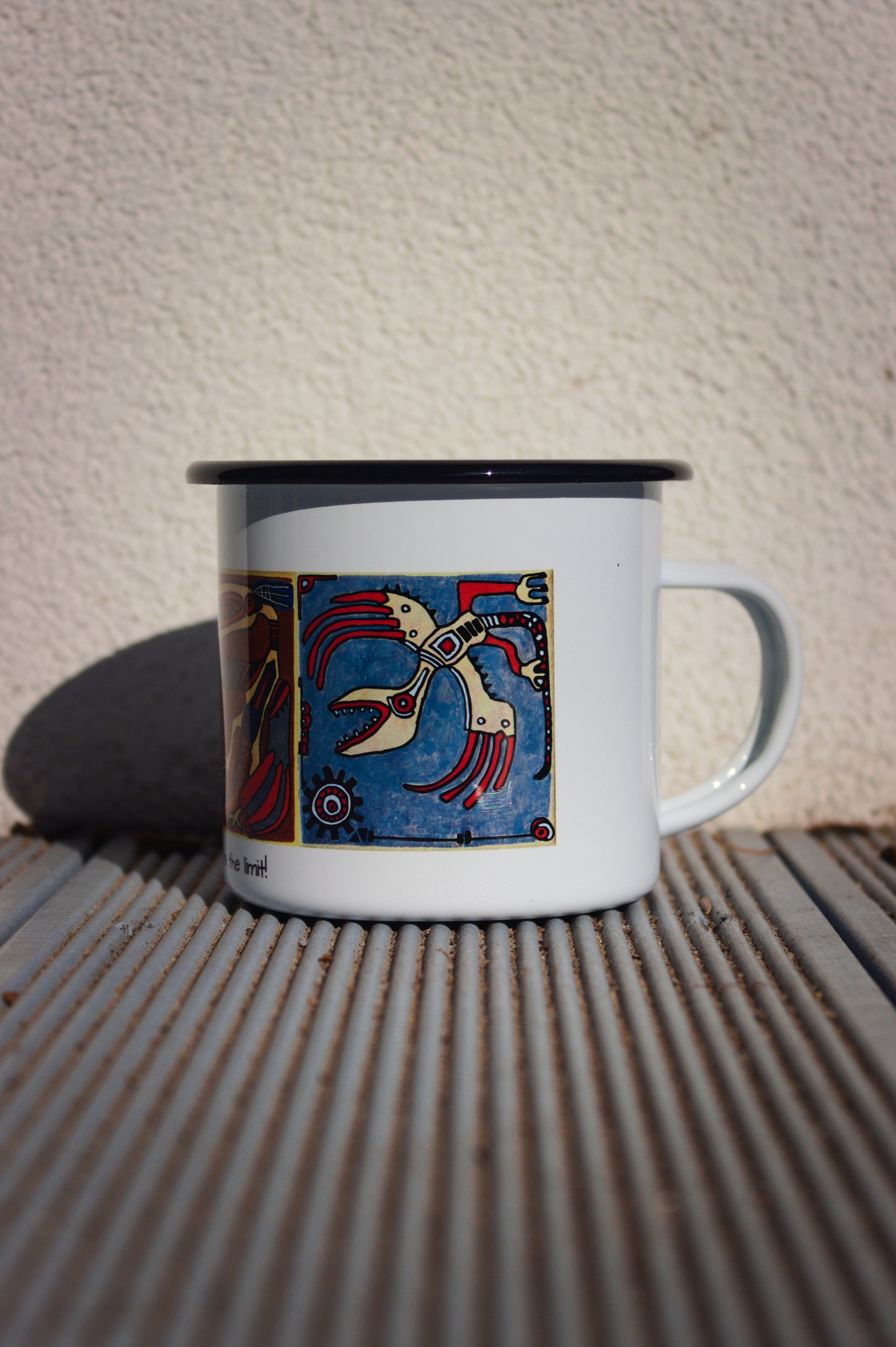 Biały kubek stalowy z czarnym rantem i motywem malarstwa kaflowego, widok na ucho i fragment wzoru.