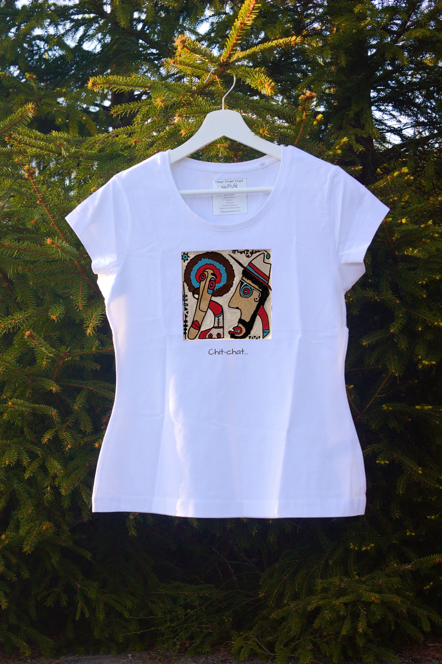 Damski t-shirt w kolorze białym zawieszony na wieszaku w otoczeniu natury. Centralnym punktem jest kolorowa, kwadratowa grafika przedstawiająca dwie postacie w profilu – jedną z kolorowym afro, drugą w kapeluszu – skierowane ku sobie. Pod grafiką umieszczono podpis: „Chit-chat…”.