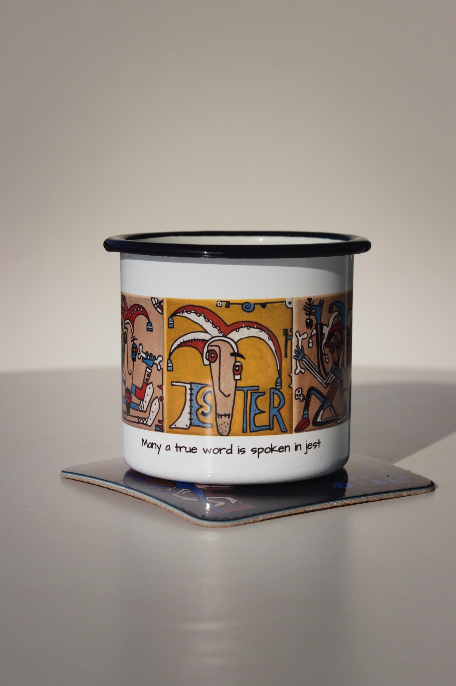 Kubek stalowy „Jester” na kolorowej podkładce z autorskim wzorem, minimalistyczne zdjęcie produktowe na jasnym tle.