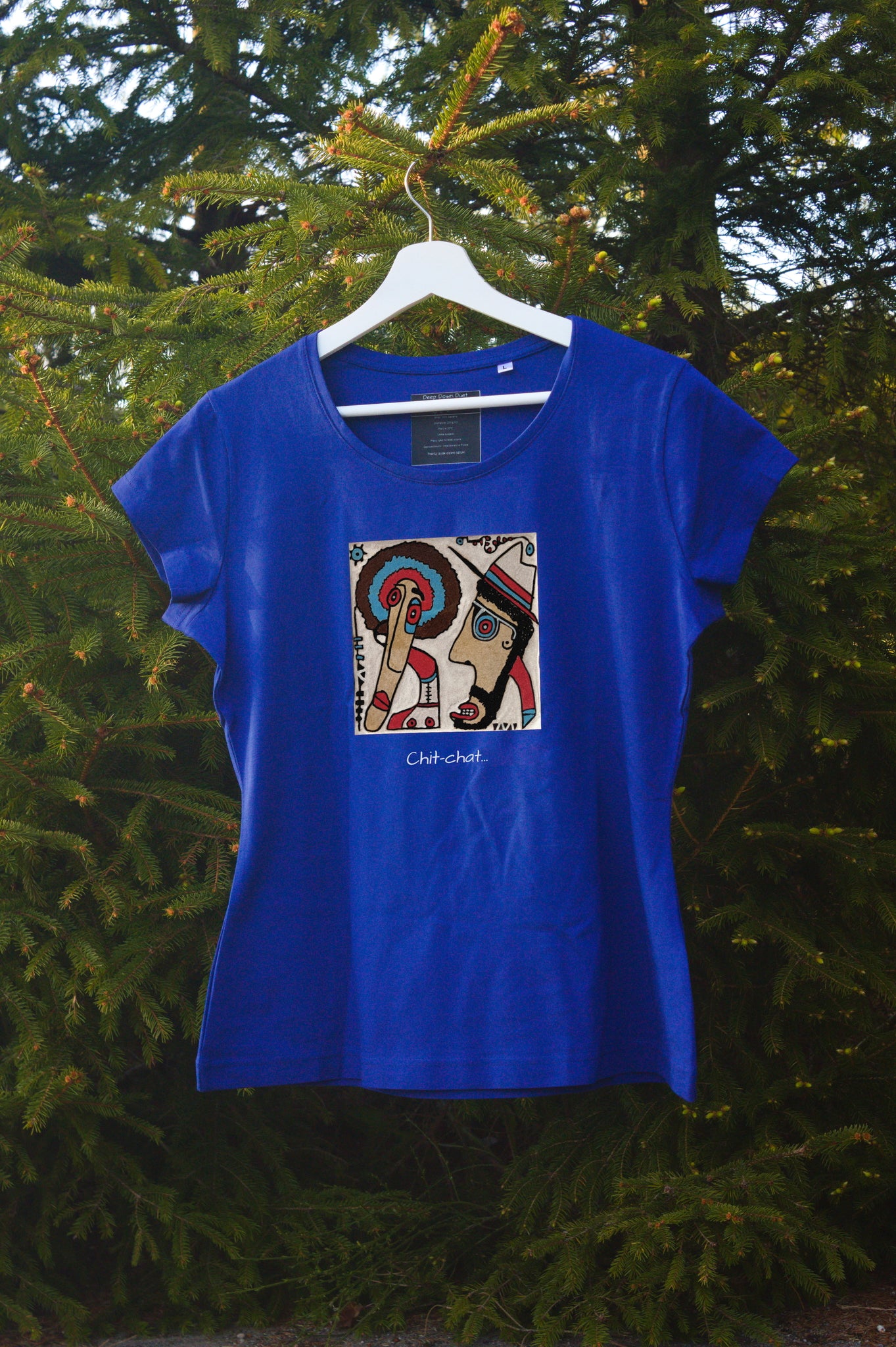 Damski t-shirt w kolorze chabrowym zawieszony na wieszaku w otoczeniu natury. Centralnym punktem jest kolorowa, kwadratowa grafika przedstawiająca dwie postacie w profilu – jedną z kolorowym afro, drugą w kapeluszu – skierowane ku sobie. Pod grafiką umieszczono podpis: „Chit-chat…”.