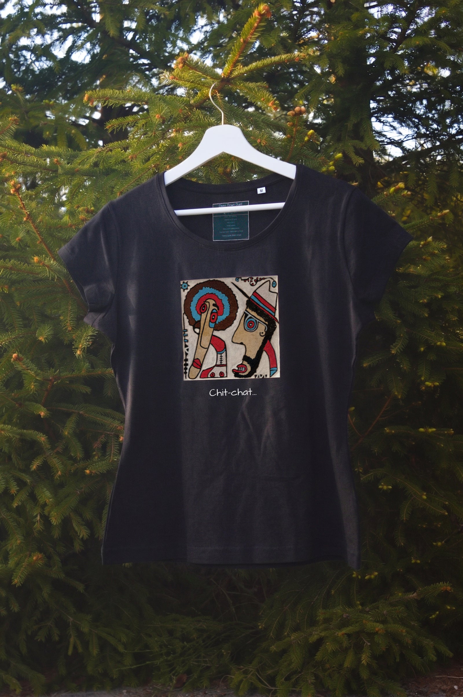 Damski t-shirt w kolorze czarnym zawieszony na wieszaku w otoczeniu natury. Centralnym punktem jest kolorowa, kwadratowa grafika przedstawiająca dwie postacie w profilu – jedną z kolorowym afro, drugą w kapeluszu – skierowane ku sobie. Pod grafiką umieszczono podpis: „Chit-chat…”.