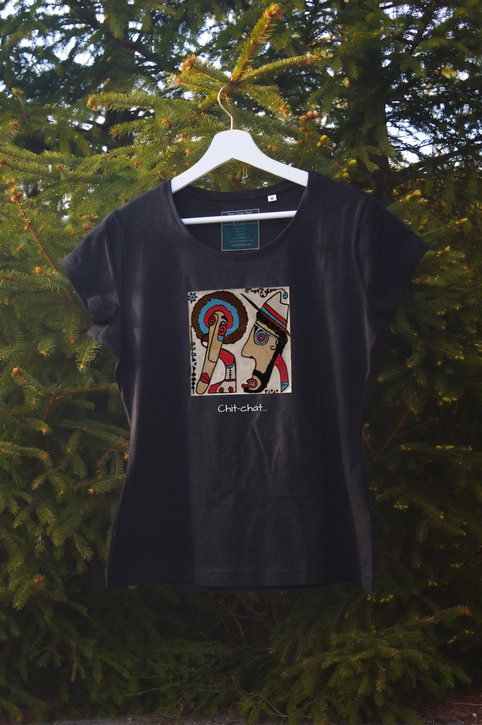 Damski t-shirt w kolorze czarnym zawieszony na wieszaku w otoczeniu natury. Centralnym punktem jest kolorowa, kwadratowa grafika przedstawiająca dwie postacie w profilu – jedną z kolorowym afro, drugą w kapeluszu – skierowane ku sobie. Pod grafiką umieszczono podpis: „Chit-chat…”.