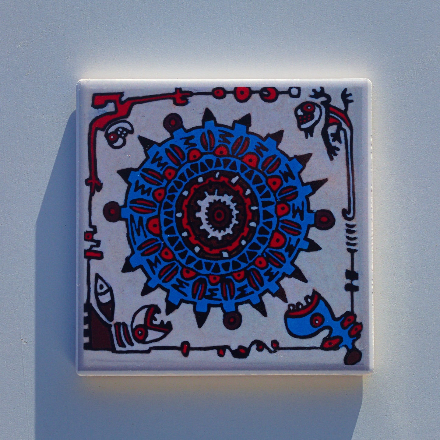 Kwadratowa ceramiczna podkładka z trwałym nadrukiem sublimacyjnym, przedstawiająca reprodukcję ręcznie malowanego projektu. Głównym motywem na białym tle jest duża, szczegółowa mandala lub tarcza przypominająca aztecki kalendarz, wykonana w odcieniach intensywnego niebieskiego, czerwieni i ciemnego brązu, z czarnymi geometrycznymi wzorami i ząbkowanymi krawędziami. W rogach podkładki znajdują się mniejsze, abstrakcyjne, plemienne stworzenia – kameleon w prawym górnym rogu oraz inne stylizowane postacie wokó