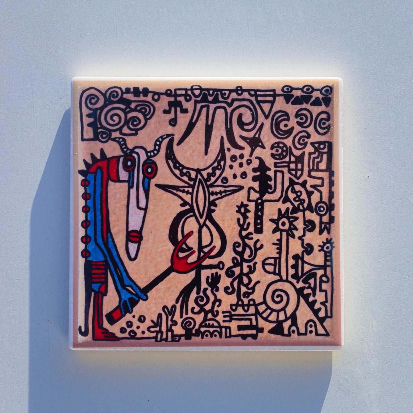 Kwadratowa ceramiczna podkładka z trwałym nadrukiem sublimacyjnym, przedstawiająca reprodukcję ręcznie malowanego projektu. Na jasnobeżowym tle znajduje się surrealistyczna, plemienna kompozycja pełna czarnych rysunków i piktogramów. Po lewej stronie widnieje antropomorficzna postać ze skręconymi rogami, długim pyskiem oraz niebiesko-czerwonym ciałem, trzymająca w dłoni czerwone widły. W centrum i po prawej stronie rozmieszczone są liczne abstrakcyjne symbole, spirale, motywy roślinne.
