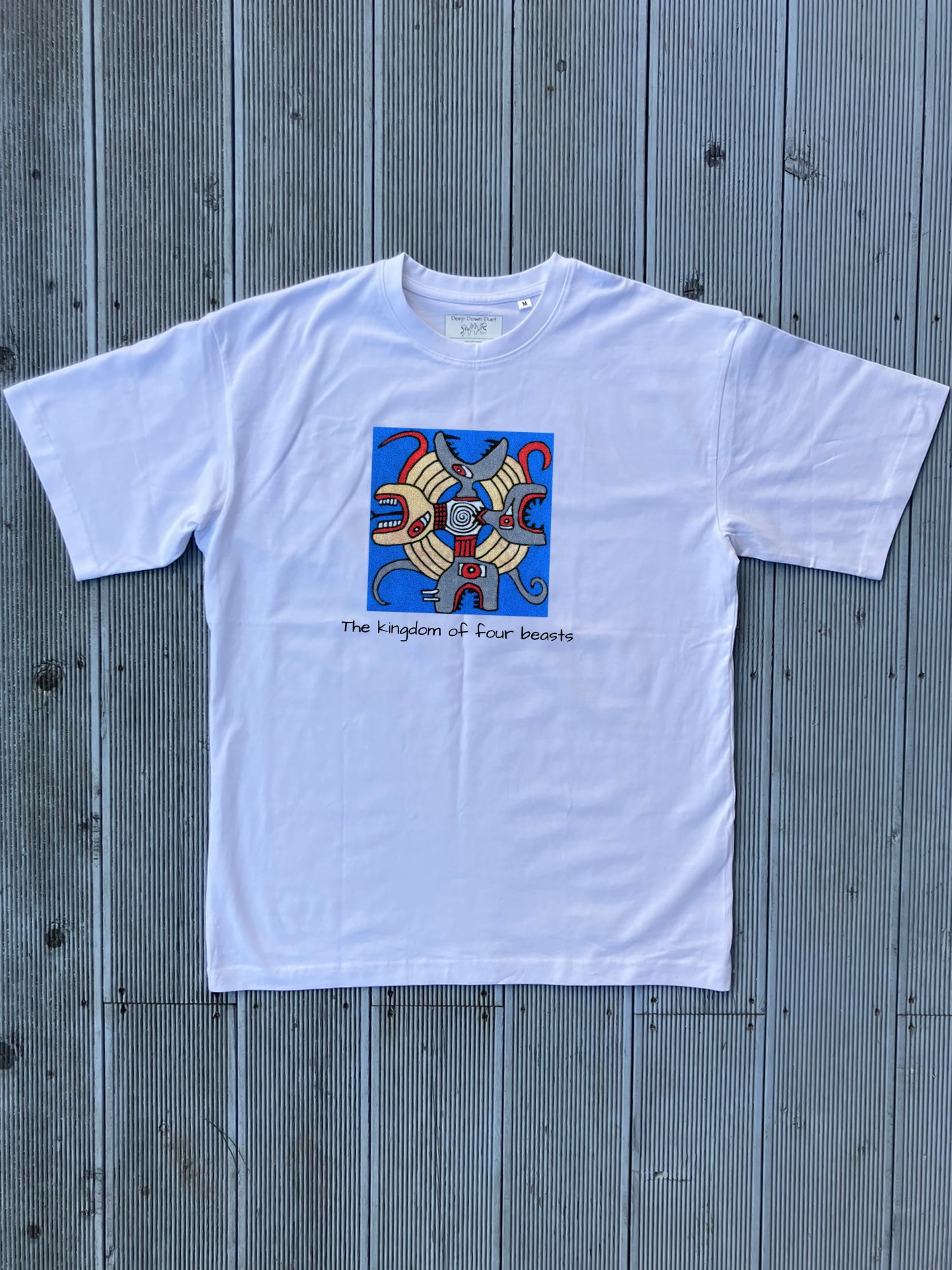 Biały t-shirt o kroju oversize z kolorowym nadrukiem kwadratowym. Grafika „The Kingdom of Four Beasts” przedstawia cztery głowy bestii z otwartymi paszczami, skierowane w cztery strony świata. Pod ilustracją znajduje się czarny napis: „The kingdom of four beasts”. Koszulka leży na tle jasnoszarych desek.
