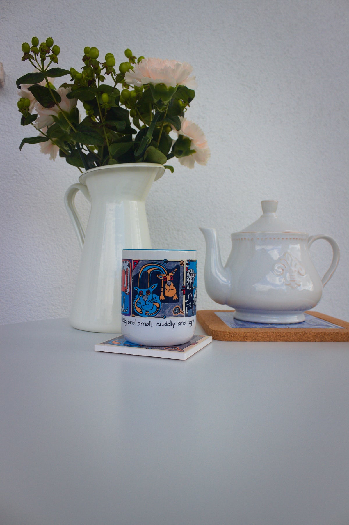 Kubek ceramiczny z grafiką kotów na stole obok wazonu z kwiatami i dzbanka herbaty.
