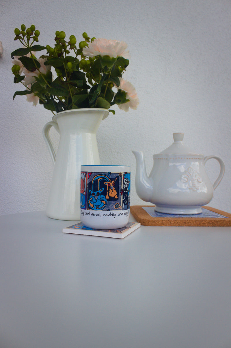 Kubek ceramiczny z grafiką kotów na stole obok wazonu z kwiatami i dzbanka herbaty.
