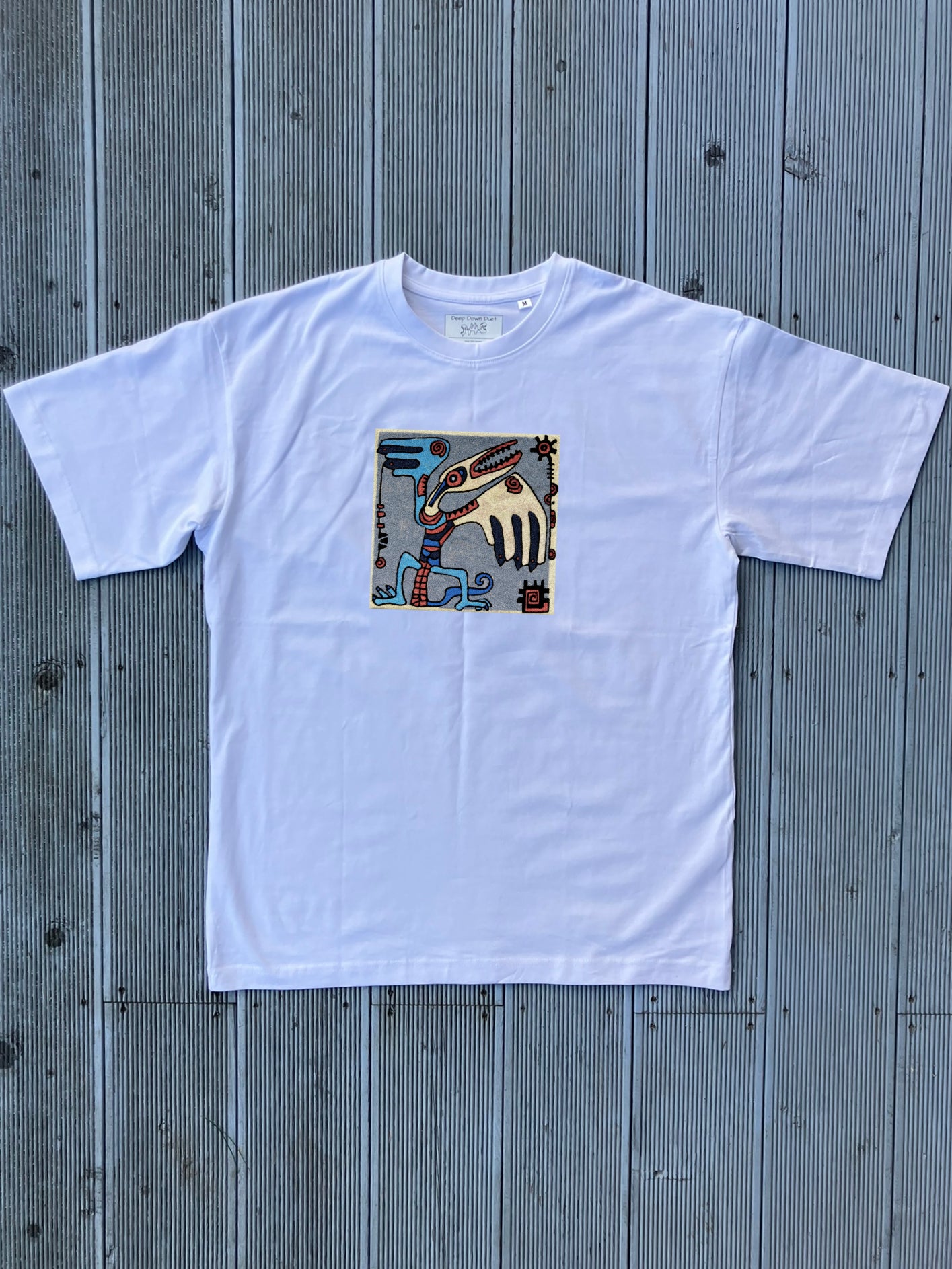 Biały t-shirt oversize z kolorową grafiką w kształcie kwadratu. Motywem przewodnim jest mityczne stworzenie z dziobem i skrzydłem, otoczone drobnymi symbolami geometrycznymi. Jasne tło koszulki mocno eksponuje detale nadruku.