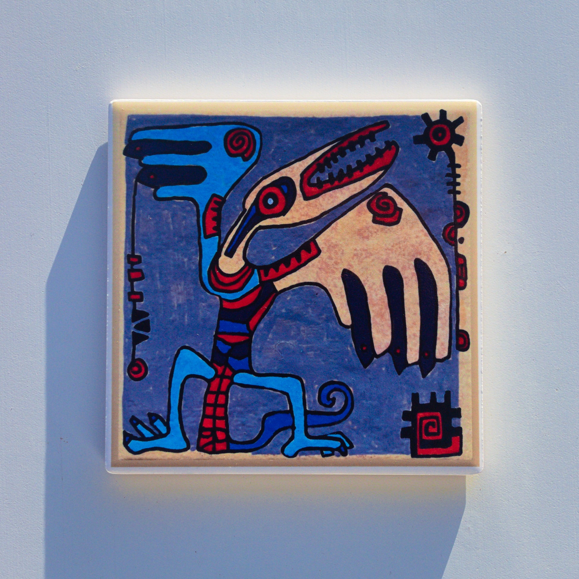 Ceramiczna podkładka z nadrukiem sublimacyjnym (reprodukcja ręcznego malunku). Abstrakcyjna, plemienna istota przypominająca ptaka lub pterodaktyla z rozpostartymi skrzydłami na szaroniebieskim tle. Kolory: błękit, beż, czerwień i czerń.