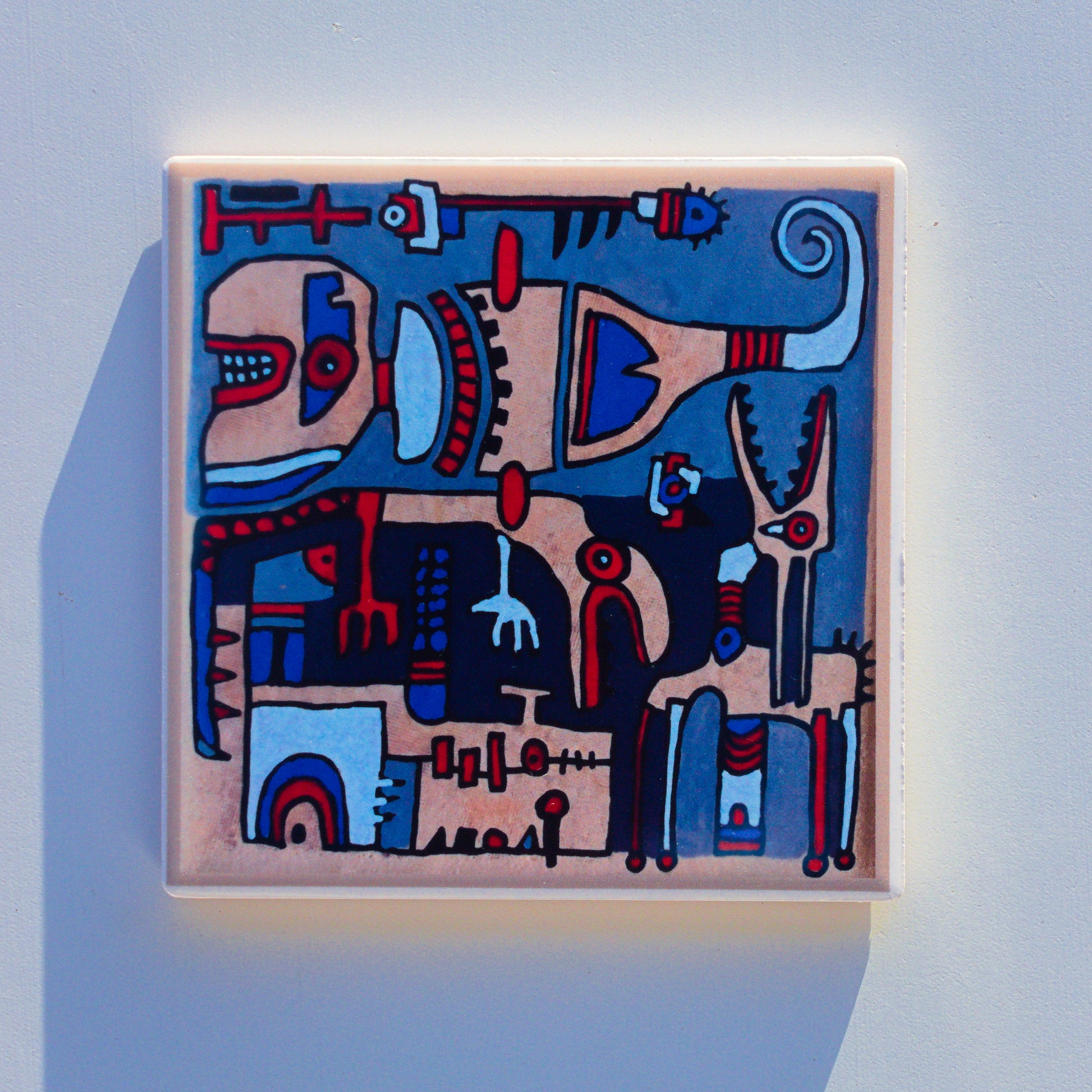 Ceramiczna podkładka z nadrukiem sublimacyjnym (reprodukcja ręcznego malunku). Złożona, abstrakcyjna kompozycja z podziałem na sekcje, przedstawiająca stylizowane postacie i stwory w klimacie plemiennym. Dominują odcienie niebieskiego, beżu i czerwieni z grubym, czarnym konturem.