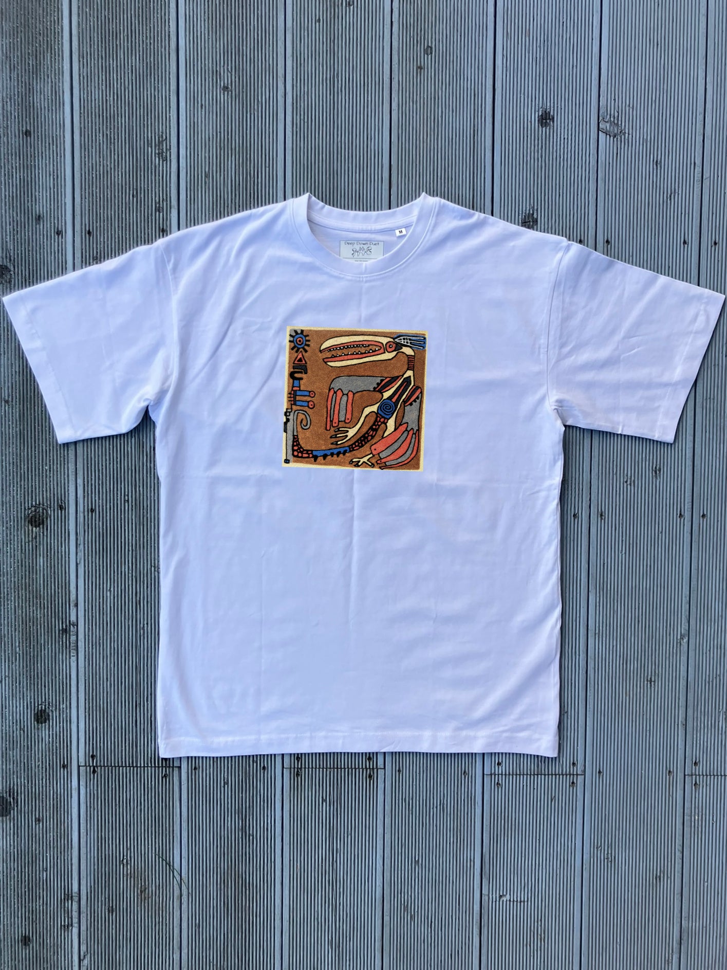 Biały t-shirt oversize z centralnie umieszczonym nadrukiem w kształcie kwadratu. Motywem jest mityczny ptak wykonany w surowym, ekspresyjnym stylu z dominacją odcieni ziemi i niebieskich akcentów. Krój koszulki jest luźny, z szerokimi rękawami.